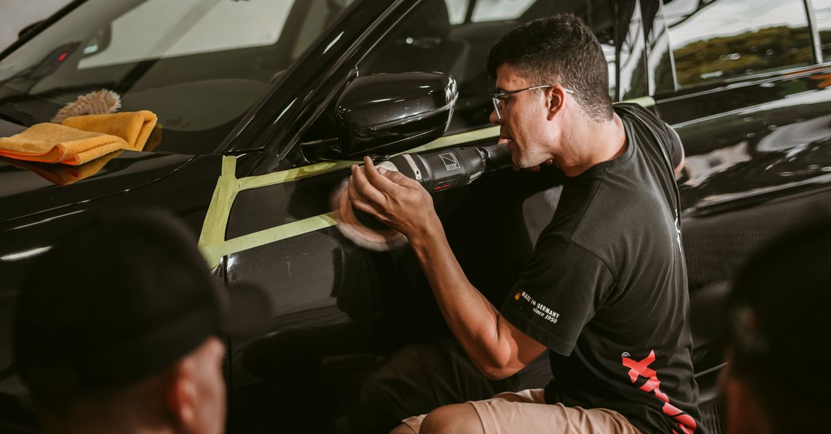 A detailer running a polisher along the door of a black car with masking tape on the trim and a microfiber towel resting on the hood