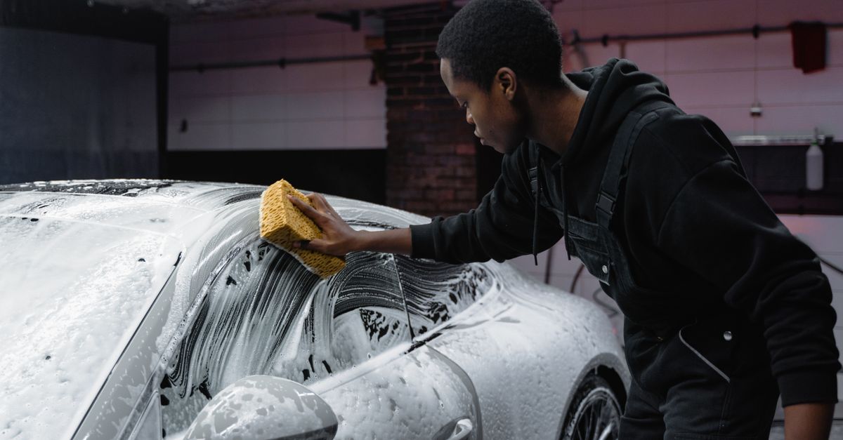 A detailer in a black jacket working a yellow sponge across the foam covered roof and windscreen of a car inside a tiled wash bay