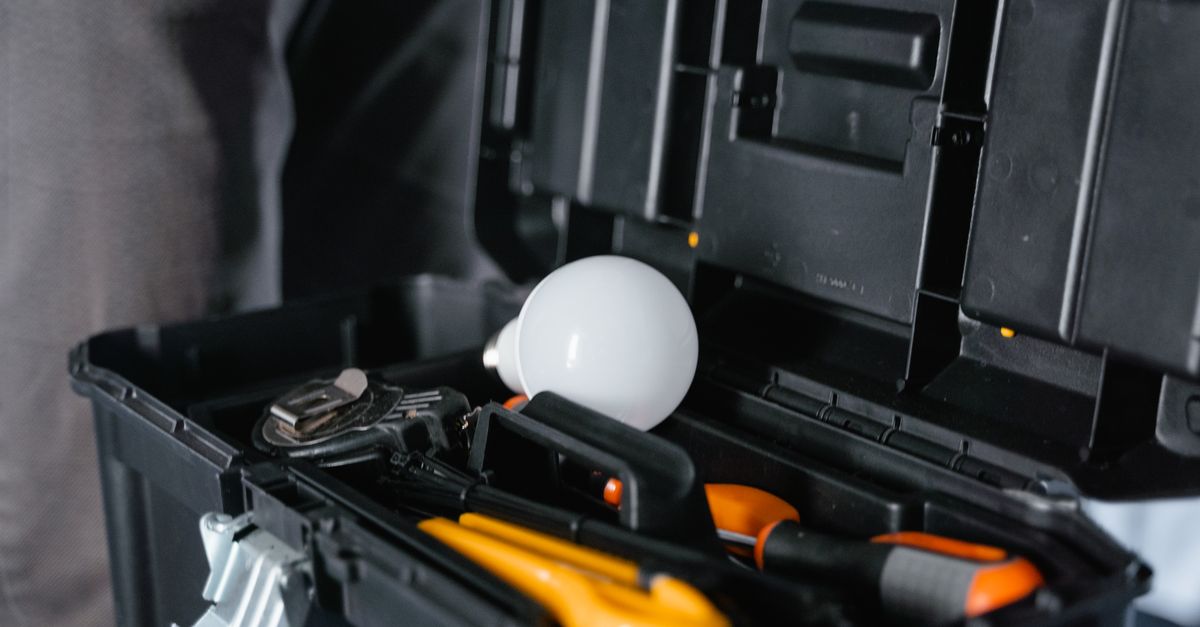 An open black toolbox holding screwdrivers, pliers and a utility knife with a light bulb resting on the tray